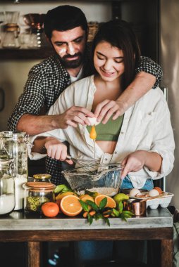 Genç mutlu çift mutfakta birlikte yemek pişiriyor. Beyaz erkek yüzü un ve esmer kadın sarılıyor, flört ediyor, gülüyor ve turunçgil pastası pişirmek için hamur malzemelerini karıştırıyor. Sevgililer Günü