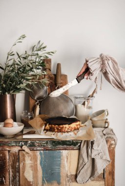 Bıçaklı ve ev yapımı San Sebastian peynirli keki kesmeye hazır kadın elleri mutfak tezgahında beyaz duvar arkasında. Zanaatkar ev yapımı tatlı konsepti