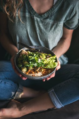 Sağlıklı akşam yemeği ya da öğle yemeği. Tişörtlü ve kot pantolonlu bir kadın elinde humus, sebze, salata, fasulye, kuskus ve elinde avokado olan vejetaryen bir kaseyle koltukta oturuyor.
