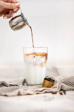 Kadın eli espresso dolduruyor ve beyaz arka planda hafif keten üzerine cam pipet içen uzun bardakta buzlu latte kahve yapıyor, seçici bir odak noktası. Kahvaltı konsepti için ideal içecek.