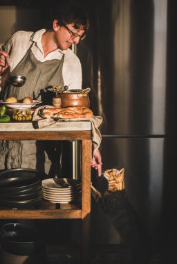 Yaşlı kadın yemek pişiriyor, tadıyor, geleneksel Türk et çorbasını sebzelerle servis ediyor ve modern mutfağın içinde kediye dokunuyor. Sonbahar konforlu gıdalar, bölgesel Orta Doğu mutfağı