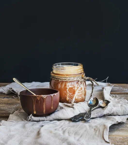 Salted caramel sauce in a rustic glass jar and brown ceramic cup on wooden desk — Stock Photo, Image