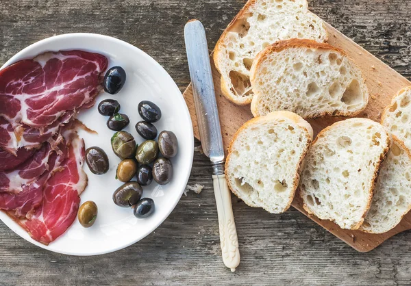 Füstölt hús, vagy sonka és olajbogyót egy fehér tábla, vintage kés, baguette szelet nyers fa háttér — Stock Fotó