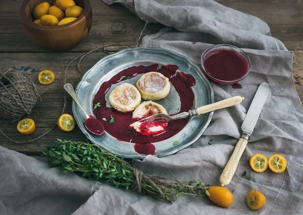 Petit déjeuner rustique. Gâteaux au fromage russe sur une plaque de métal vintage avec confiture d'airelles, kumquats frais, thym, corde de décoration et vaisselle ancienne sur un bureau en bois rugueux — Photo