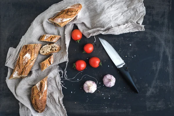 Французский багет разрезанный на куски, помидоры черри, чеснок и кухонный нож на темном фоне гранжа — стоковое фото