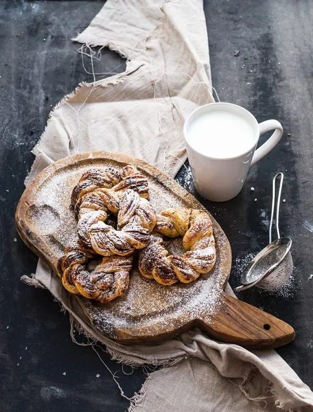 Pães de canela com açúcar em pó em tábua de madeira rústica, caneca de leite, superfície grunge escuro — Fotografia de Stock