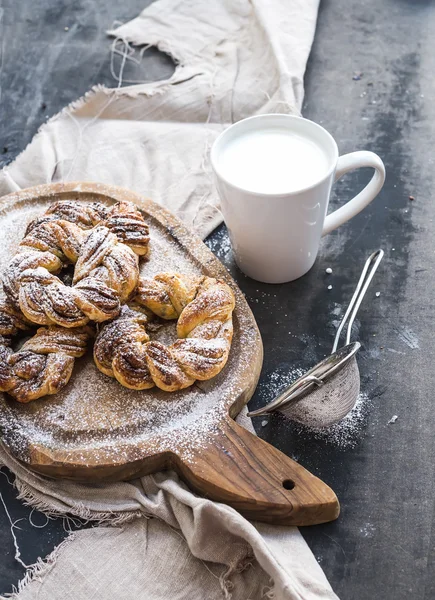 Zimtbrötchen mit Zuckerpuder auf rustikalem Holzbrett, Becher Milch, dunkle Grunge-Oberfläche — Stockfoto