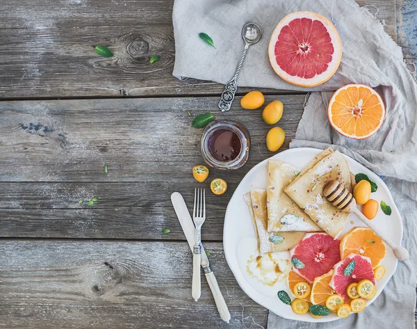 Spring vitamin breakfast set. Thin crepes or pancakes with fresh grapefruit, orange, kumquat, honey, cream and mint leaves over a rustic wood background — Stock Photo, Image