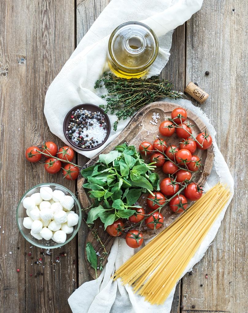 Ingredients for cooking pasta. Spaghetti, basil, cherry-tomatoes, mozarella, olive oil, thyme, salt, spices on rustic chopping board over old wood background