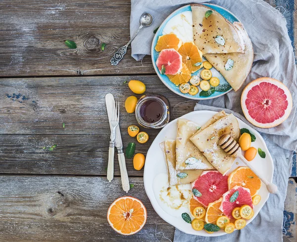 Spring vitamin breakfast set. Thin crepes or pancakes with fresh grapefruit, orange, kumquat, honey, cream and mint leaves over a rustic wood background