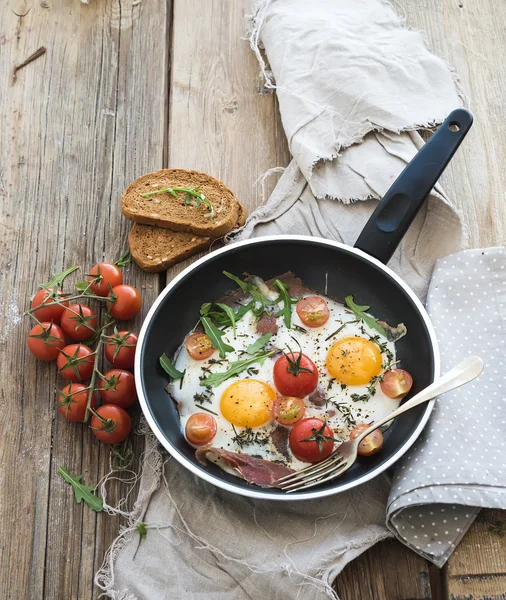 Pan of fried eggs, bacon and cherry-tomatoes with bread on rustic wood table surface — Stock Photo, Image