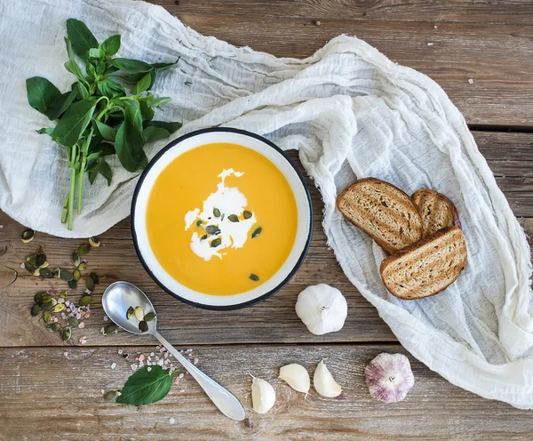 Pumpkin soup with cream, seeds, bread and fresh basil in metal plate on rustic wood background. — Stock Photo, Image