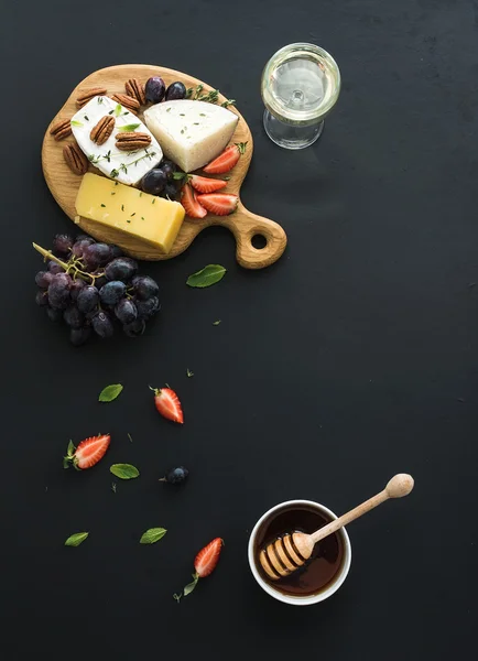 Sajt válogatás előétel, vagy nyafogás snack készlet. Különböző sajt, szőlő, pekándió, eper és méz kerek fából készült táblán át fekete hátteret, felülnézet — Stock Fotó