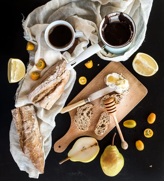 stock image Breakfast set: pot or cezve of coffee, cup on kitchen towel, kumquats, pears, grapefruit, baguette slices with butter cream and honeyl, rustic wooden board over  black backdrop