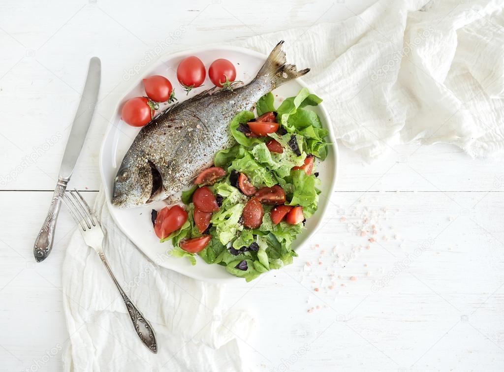 Cooked sea bream fish with fresh vegetable salad on ceramic plate over white rustic wooden backdrop, top view