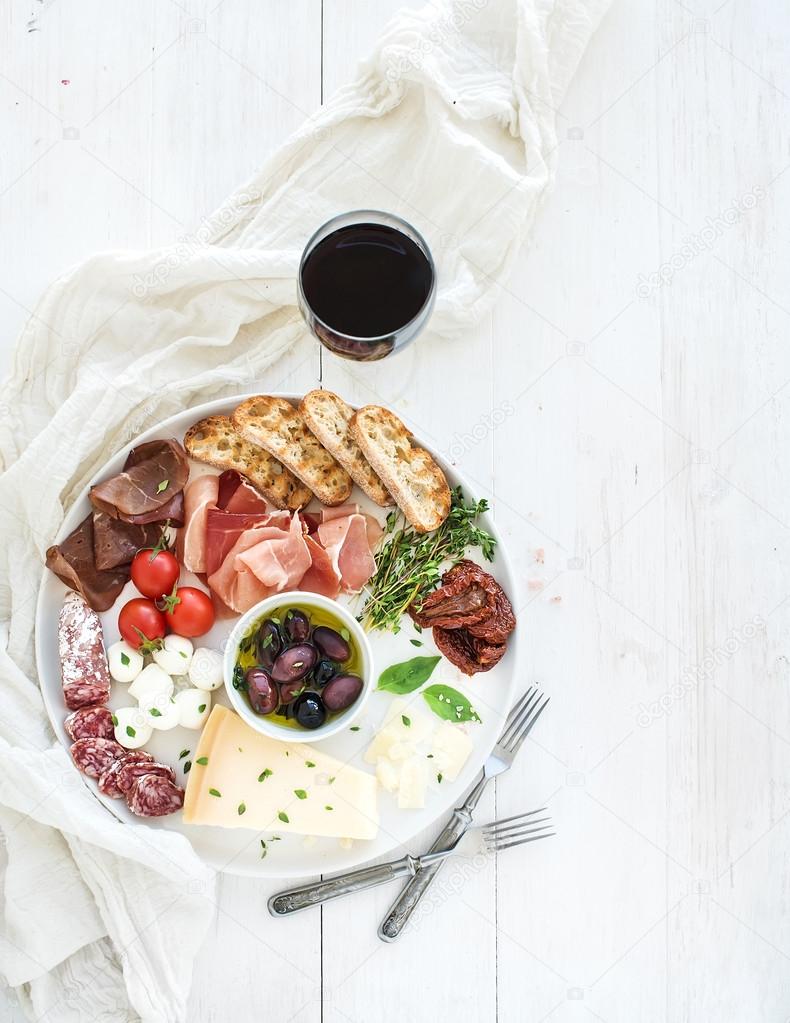 Wine appetizer set. Cherry-tomatoes, parmesan cheese, meat variety, bread slices, dried tomatoes, olives and basil on round ceramic plate over white wood backdrop