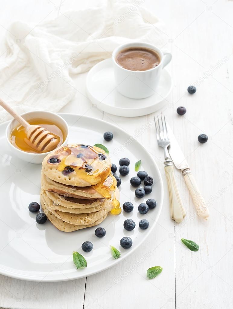 Breakfast set. Blueberry pancakes with fresh berries, honey, mint leaves and cup of coffee over white wooden background