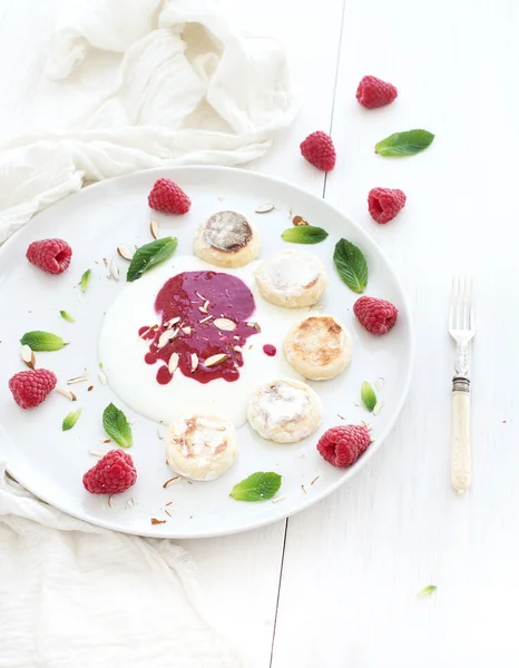 Colazione rustica. Torte russe di fiocchi di latte su un piatto di ceramica con marmellata di bacche, mandorle, lamponi freschi e menta su sfondo di legno bianco, vista dall'alto — Foto Stock