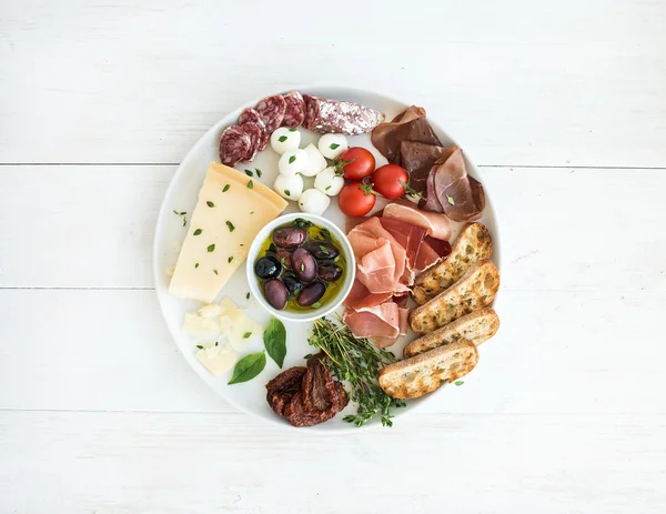 Set de aperitivos de vino. Tomates cereza, queso parmesano, variedad de carne, rebanadas de pan, tomates secos, aceitunas y albahaca en plato de cerámica redonda sobre fondo de madera blanca —  Fotos de Stock