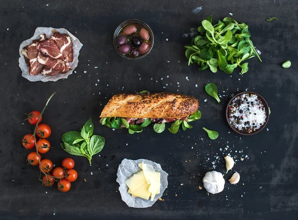 Ingredients for sandwich with smoked meat, baguette, basil, arugula, olives, cherry-tomatoes, parmesan cheese, garlic and spices over black grunge background — Stock Image Ingredients for sandwich with smoked meat, baguette, basil, arugula, olives, cherry-tomatoes, parmesan cheese, garlic and spices over black grunge background Stock Photo