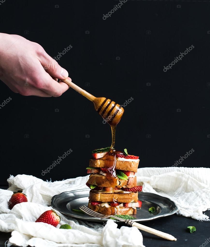 French toasts tower with strawberry, cream cheese and mint, hand is pouring honey over top of it