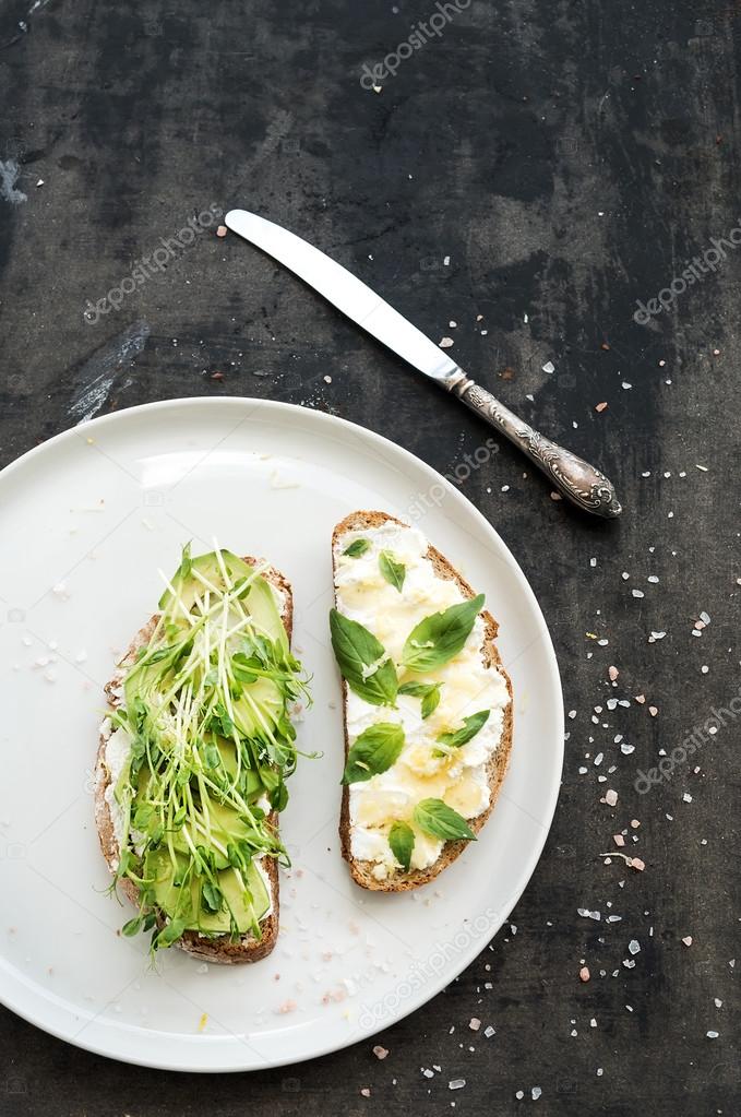 Avocado, ricotta, basil and sprout sandwiches on white ceramic plate over dark grunge backdrop, top view