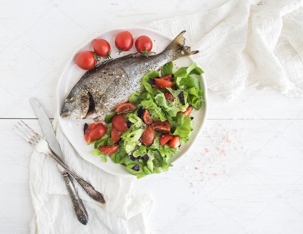 Cooked sea bream fish with fresh vegetable salad on ceramic plate over white rustic wooden backdrop, top view