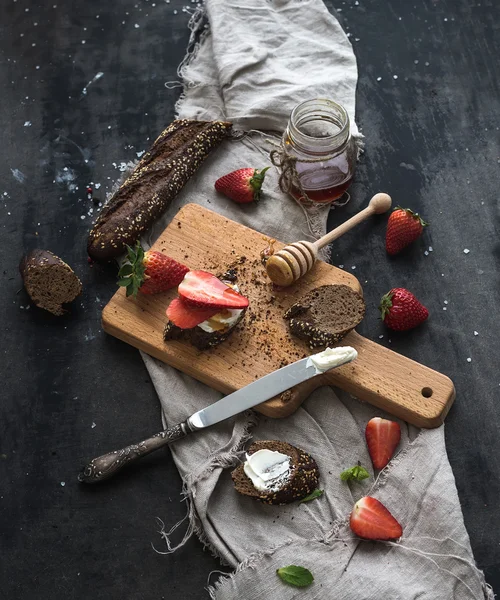 Colazione pronta. Crostini alla baguette nera con fragole fresche, miele e mascarpone sulla superficie scura della tavola grunge — Foto Stock