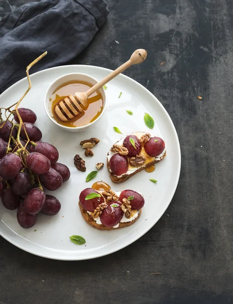 Colazione pronta. Panini dolci con ricotta, uva, noci e miele su piastra in ceramica bianca su fondo grunge scuro — Foto Stock