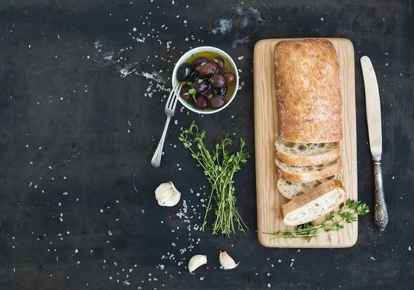 Italian ciabatta bread cut in slices on wooden chopping board with herbs, garlic and olives over dark grunge backdrop, copy space