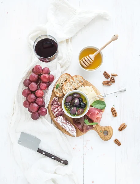 Wine appetizer set. Glass of red, grapes, parmesan cheese, meat variety, bread slices, pecan nuts, honey, olives and basil on rustic wooden board over white backdrop — Stock Photo, Image