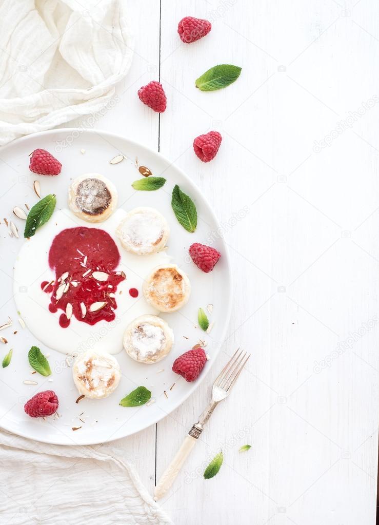 Rustic breakfast set. Russian cottage cheese cakes on ceramic plate with berry jam, almonds, fresh raspberries and mint over white wooden backdrop, top view