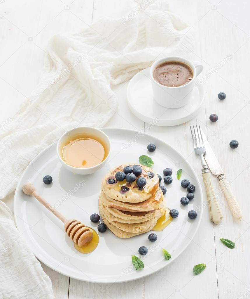 Breakfast set. Blueberry pancakes with fresh berries, honey, mint leaves and cup of coffee over white wooden background