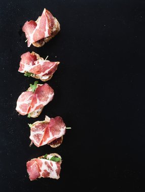 Bruschettas with prosciutto smoked meat, dried tomatoes and arugula over  black backdrop, top view