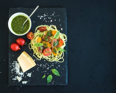 Spaghetti with pesto sauce, roasted cherry-tomatoes, fresh basil and parmesan cheese on black stone serving board over dark grunge backdrop. Top view