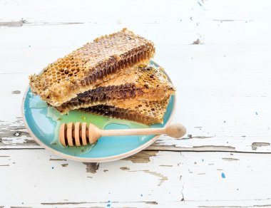 Honeycomb with honey dipper on blue ceramic plate over rustic white wooden table.