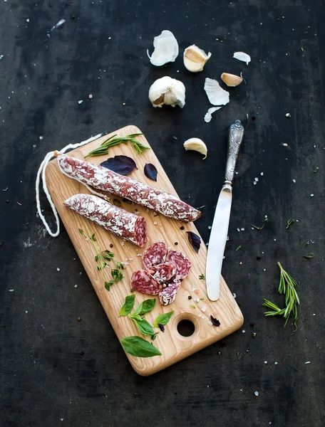 Meat gourmet snack. Salami, garlic, baguette and herbs on rustic wooden board over dark grunge backdrop — Stock Photo, Image