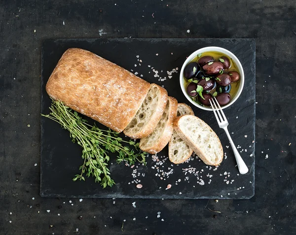 Pão ciabatta italiano cortado em fatias em tábua de corte de madeira com ervas, alho e azeitonas sobre fundo grunge escuro, espaço de cópia — Fotografia de Stock