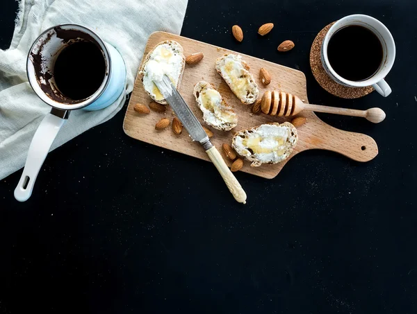 Frokostsett: kanne eller kanne av kaffe, beger, kjøkkenhåndkle, bagettskiver med smørkrem, mandler og honning på rustikt trebord over svart bakteppe – stockfoto