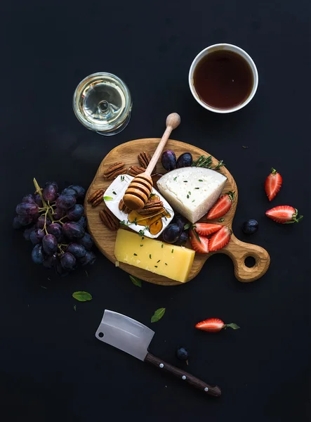 Selección de aperitivo de queso o conjunto de aperitivos whine. Variedad de quesos, uvas, nueces de nuez, fresa y miel sobre tabla redonda de madera sobre fondo negro — Foto de Stock