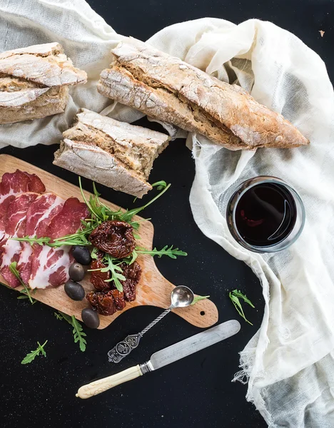 Conjunto de aperitivos de vinho: louça de jantar vintage, baguete francesa quebrada em pedaços, tomates secos, azeitonas, carne defumada e rúcula em tábua de madeira rústica sobre fundo escuro — Fotografia de Stock