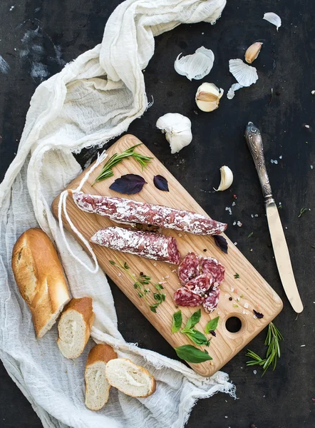 Hús ínyenc snack. Szalámi, fokhagyma, baguette és gyógynövények a rusztikus, fából készült fedélzet fölött sötét grunge hátteret — Stock Fotó