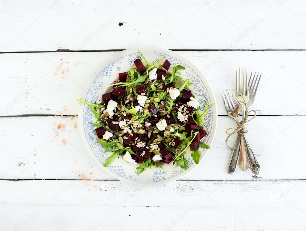 Beetroot salad with arugula, feta cheese, red salt and pumpkin seeds in vintage plate over white rustic wooden background, top view