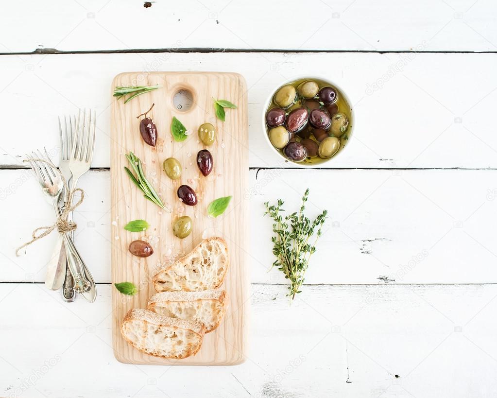 Mediterranean olives with herbs and ciabatta slices on rustic wooden board  over white background, top view