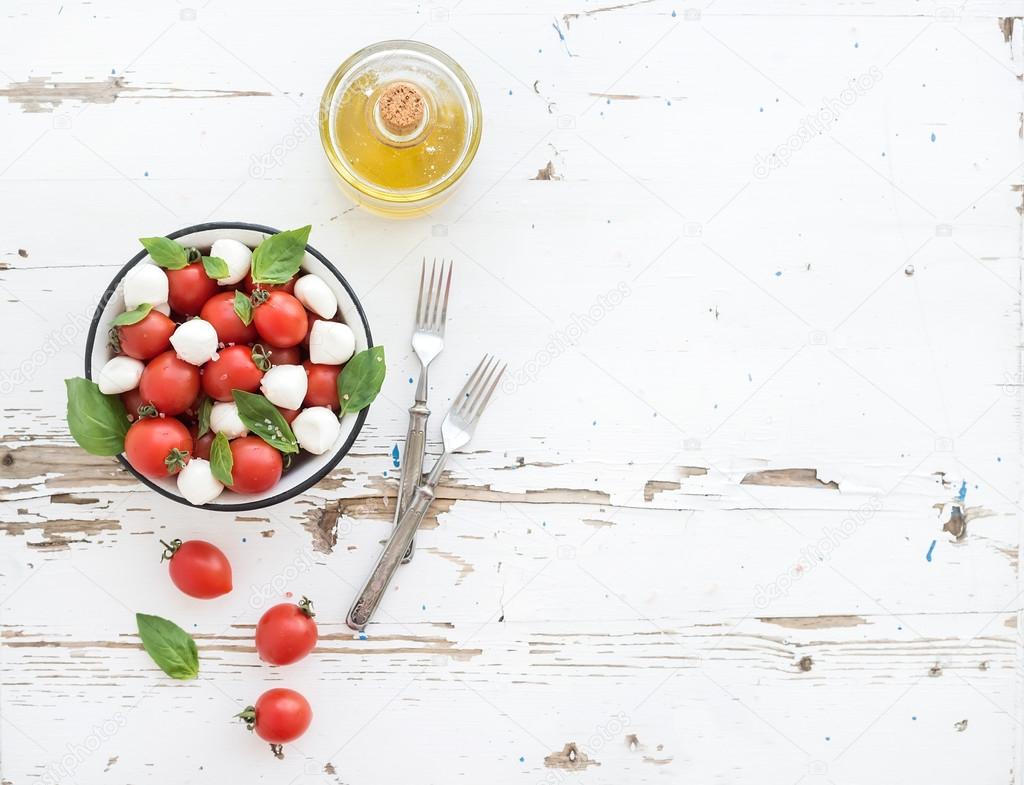 Caprese salad: cherry-tomatoes and mozzarella in metal bowl with olive oil on rustic white wooden backdrop, top view,