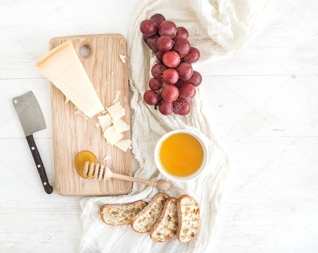 Parmesan cheese with grapes, honey and bread slices on wooden chopping board over rustic white background. Top view