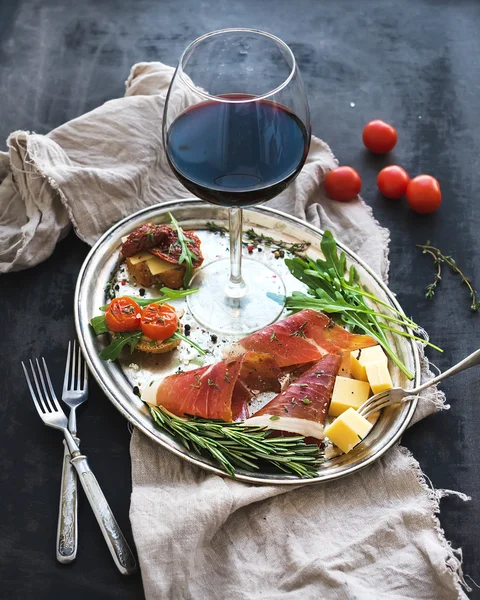 Wine appetizer set. Glass of red wine, vintage dinnerware, brushetta with cherry, dried tomatoes, arugula, parmesan, smoked meat on silver tray over rustic grunge surface — Stock Photo, Image