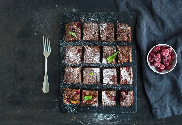 Cuadrados de brownie de chocolate y frambuesa en tablero de piedra negro sobre fondo grunge oscuro — Foto de Stock