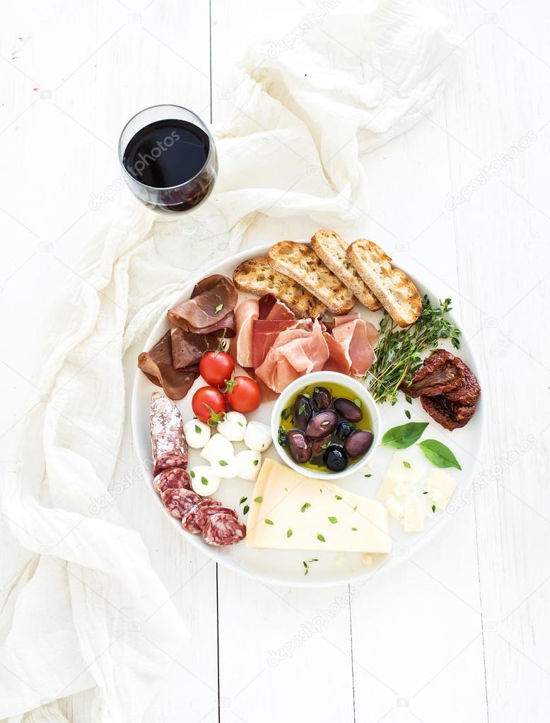 Wine appetizer set. Cherry-tomatoes, parmesan cheese, meat variety, bread slices, dried tomatoes, olives and basil on round ceramic plate over white wood backdrop