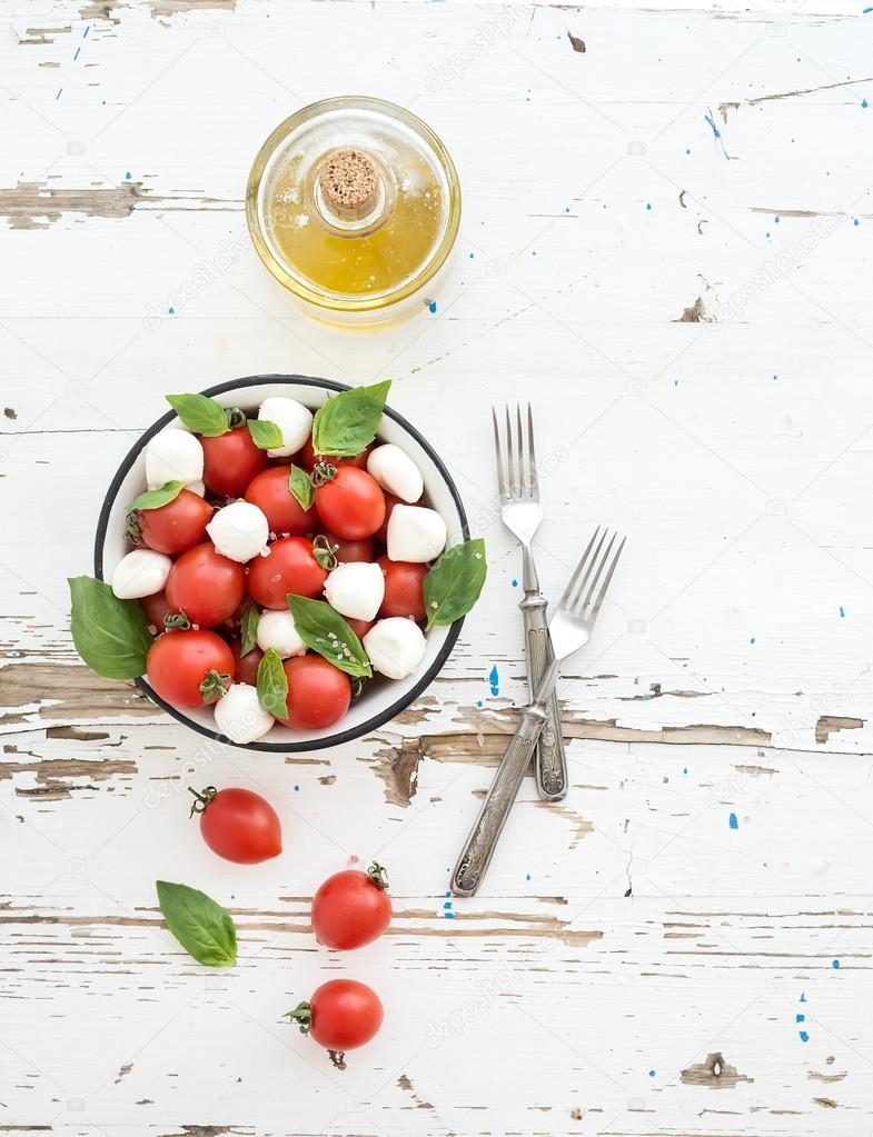 Caprese salad: cherry-tomatoes and mozzarella in metal bowl with olive oil on rustic white wooden backdrop, top view,